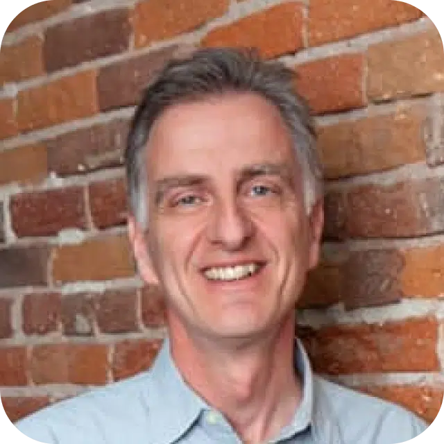 Smiling professional man in a casual shirt against a brick wall backdrop.