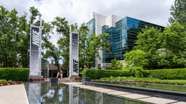 A modern, glass-paneled building with lush greenery and a reflective water feature in front. Tall white banners with "NIKE" text stand on either side. The setting is bright and sunny with a partly cloudy sky.