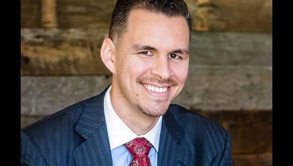 A man in a dark pinstripe suit, white shirt, and red patterned tie smiles at the camera. He has short dark hair and a trimmed goatee, with a wooden textured background behind him.