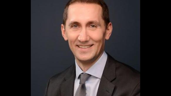 A man wearing a dark suit, light blue shirt, and patterned tie smiles at the camera against a plain dark background.