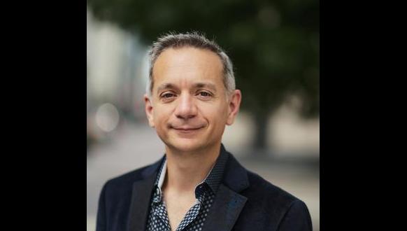 A middle-aged man with short gray hair and a slight smile, wearing a dark blazer over a patterned shirt, stands outdoors with a blurred background of greenery and buildings.
