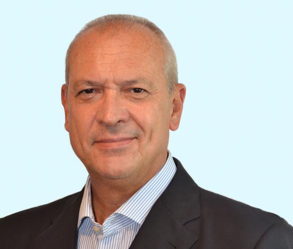 A middle-aged man with short gray hair wearing a dark suit jacket and a light blue striped shirt, smiling slightly, posed against a light blue background.