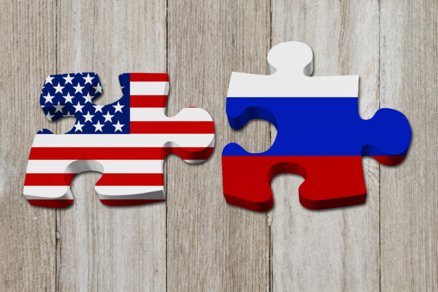Two puzzle pieces on a wooden surface, one with the American flag design and the other with the Russian flag design, placed next to each other but not connected.