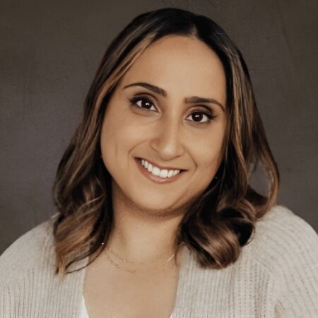 A woman with shoulder-length brown hair and dark eyes smiles at the camera. She is wearing a light-colored sweater over a white top, with a neutral, dark background.