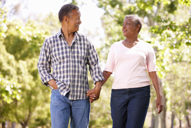 An African American couple walks hand-in-hand outdoors, smiling at each other amongst blurred green trees.